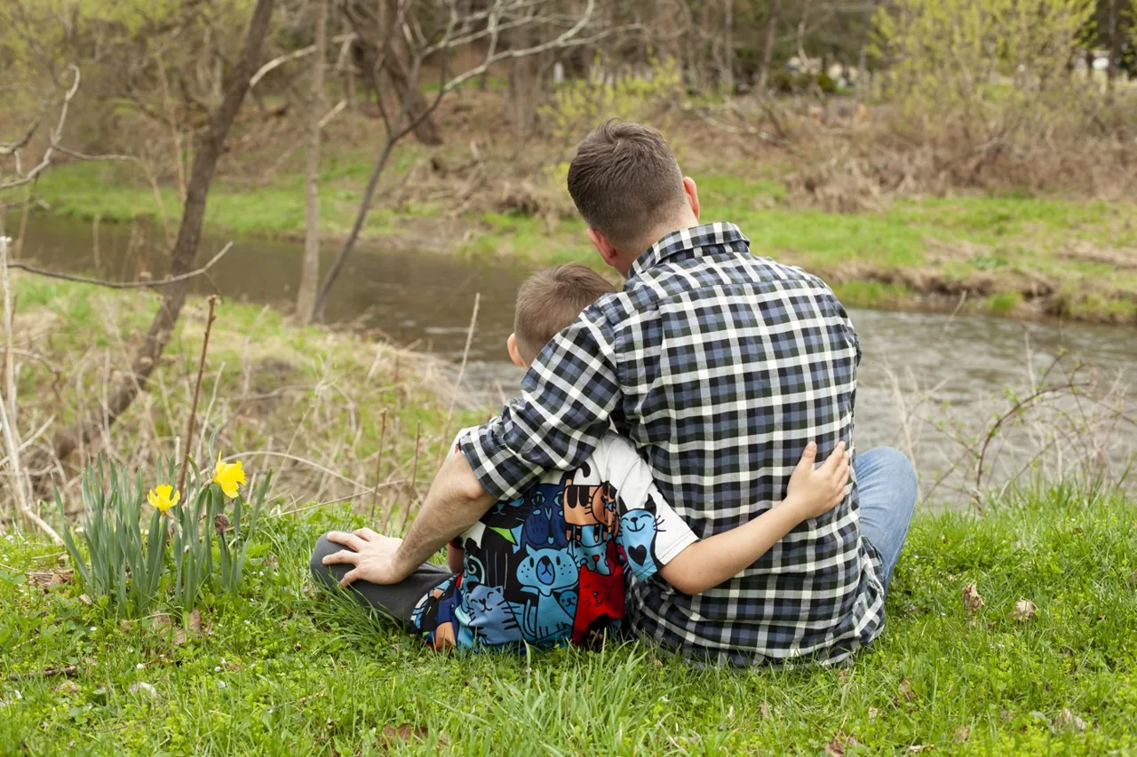 Jason and a child seated together beside a creek, with their arms around each other.